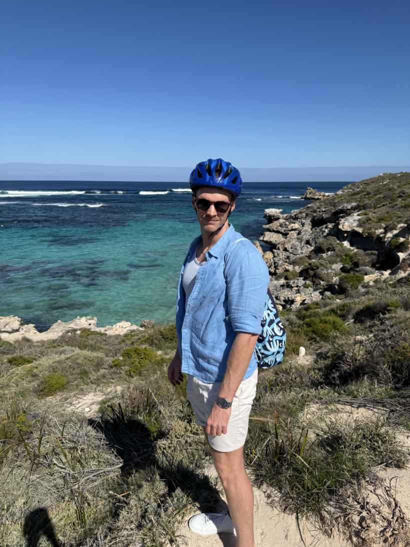Krzysztof z Ozland Travel w kasku rowerowym przy plaży na Rottnest Island, z widokiem na zatokę i turkusową wodę Oceanu Indyjskiego.