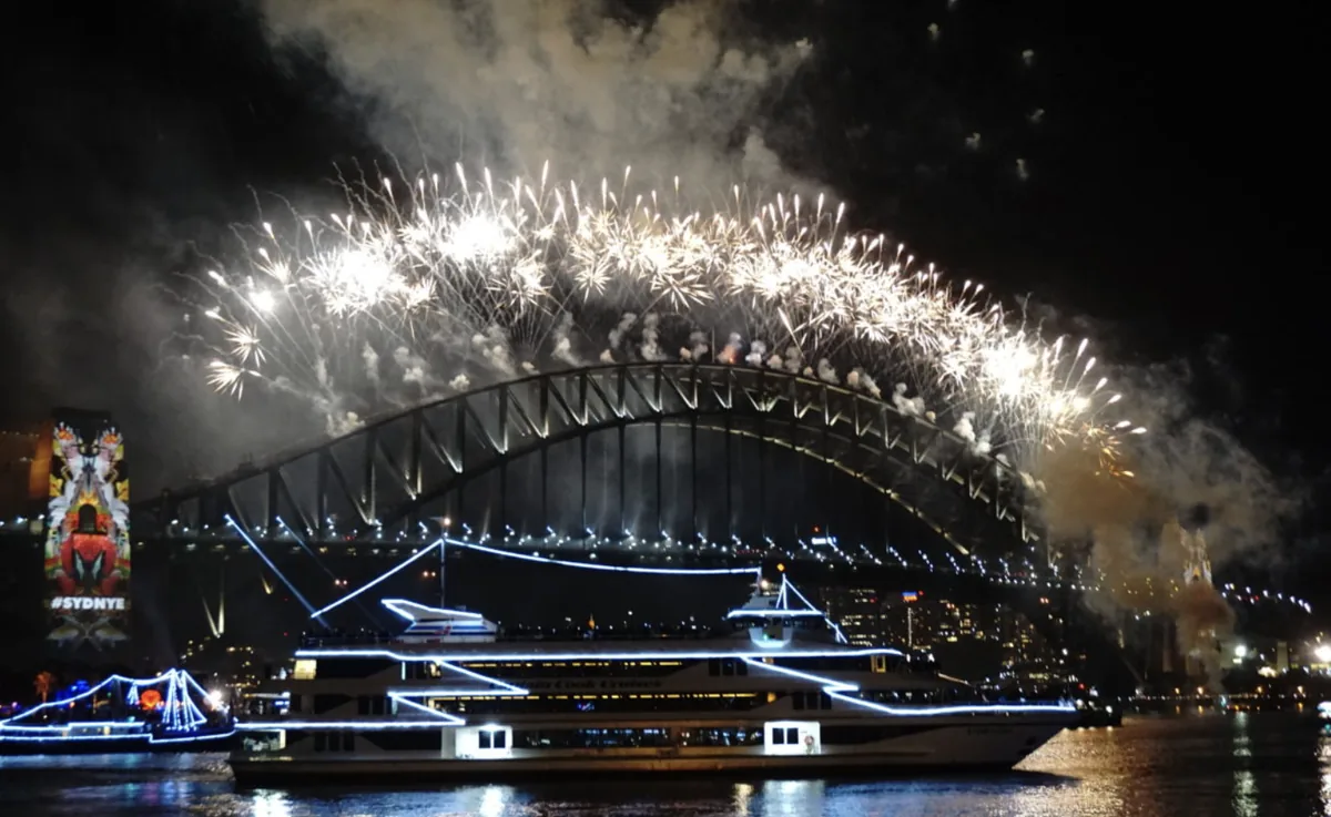 Oświetlony statek wycieczkowy podczas rejsu sylwestrowego w Sydney, nad nim fajerwerki na Harbour Bridge